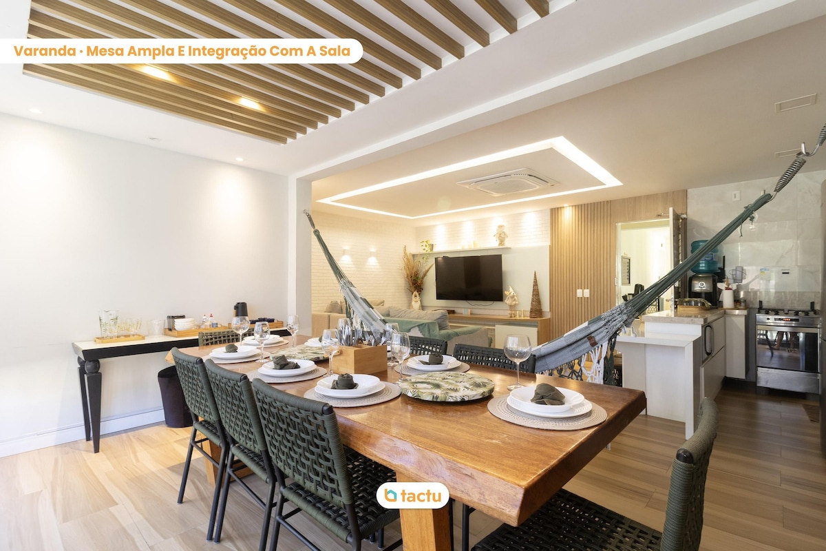 A spacious dining area is highlighted by a large wooden table set for guests. Decorative tableware is arranged thoughtfully. A hammock adds a relaxed touch to the space, which connects seamlessly to the adjacent kitchen and living area, reflecting an open-concept design.