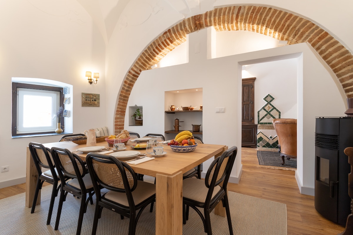 A spacious dining area is centered around a wooden table surrounded by chairs. Decorative elements include a bowl of fresh fruit and dishes. Brick arches frame the room, and a pellet stove is visible, contributing to a cozy ambiance.