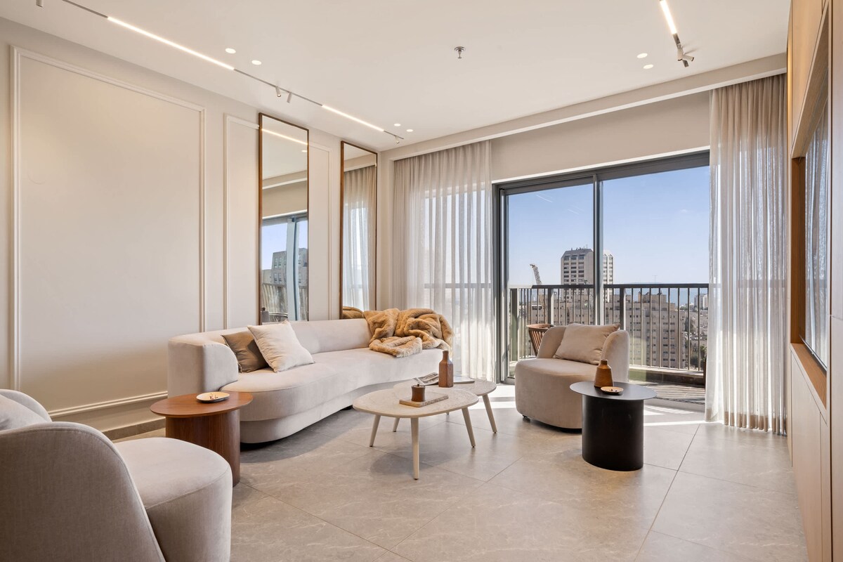 The spacious living area features a modern sofa and two accent chairs, arranged around a circular coffee table. Large windows provide ample natural light and a clear view of the city skyline, while light-colored curtains enhance the open ambiance.