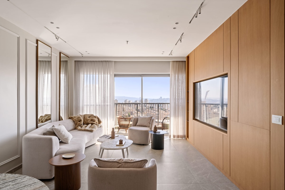 A modern living room is showcased, featuring a spacious sofa and two soft armchairs. A round coffee table is placed at the center, complemented by large windows that provide a panoramic view of the city. Elegant curtains gently filter natural light into the space.
