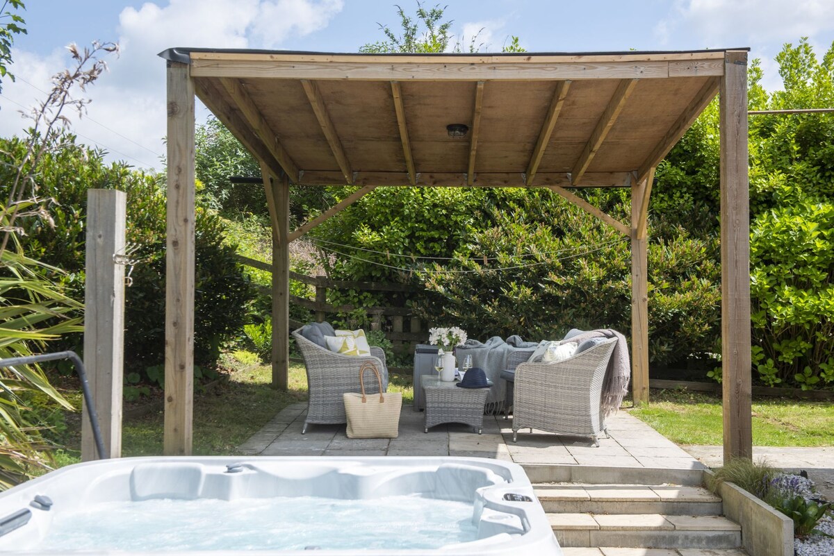 A shaded outdoor seating area is shown beneath a wooden pergola, furnished with comfortable chairs and a small table. Lush greenery surrounds the space, creating a secluded atmosphere. A bubbling hot tub is positioned in the foreground, inviting relaxation amidst nature.
