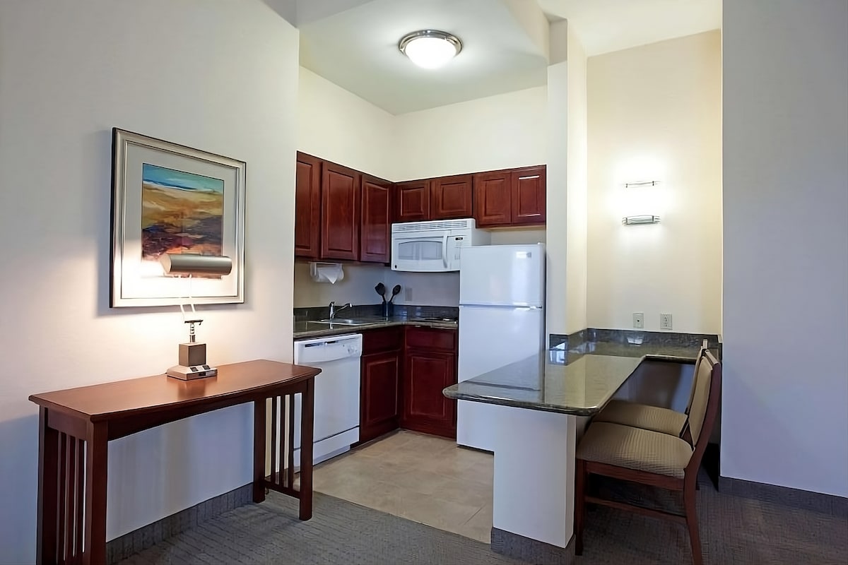 A well-equipped kitchen area features wooden cabinetry and modern appliances, including a full-sized refrigerator, microwave, and dishwasher. A dining table with a sleek lamp occupies one corner, while a granite countertop supports additional workspace, creating a functional and inviting layout.