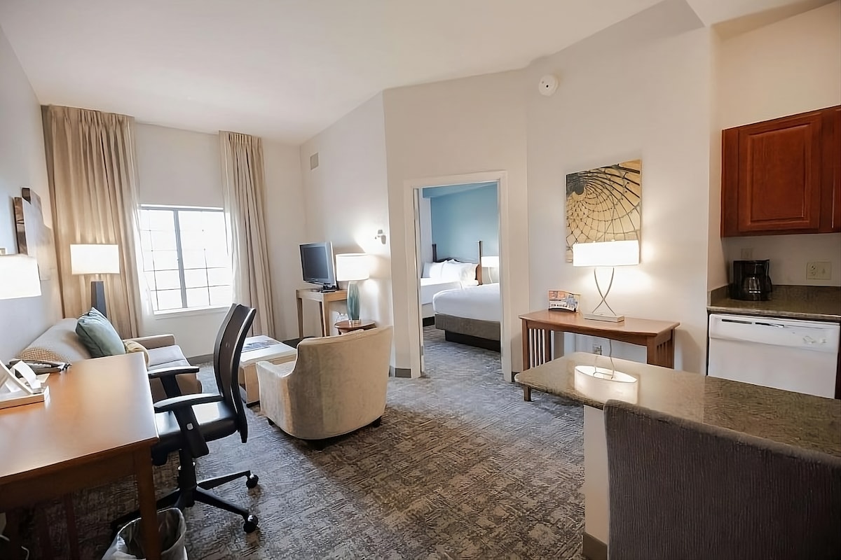 A spacious one-bedroom suite is showcased, featuring two queen beds in a separated area. A comfortable living space includes a sofa chair, desk, and flat-screen TV. A fully-equipped kitchen area is visible with cabinetry and appliances, while natural light filters through a large window.