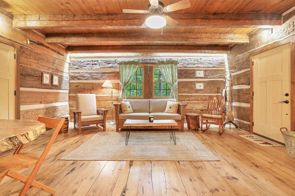 A rustic living area features a cozy sofa against log walls, complemented by wooden furniture and a woven area rug. Natural light enters through double windows adorned with sheer curtains, highlighting the warm tones of the wooden flooring and inviting seating arrangements.