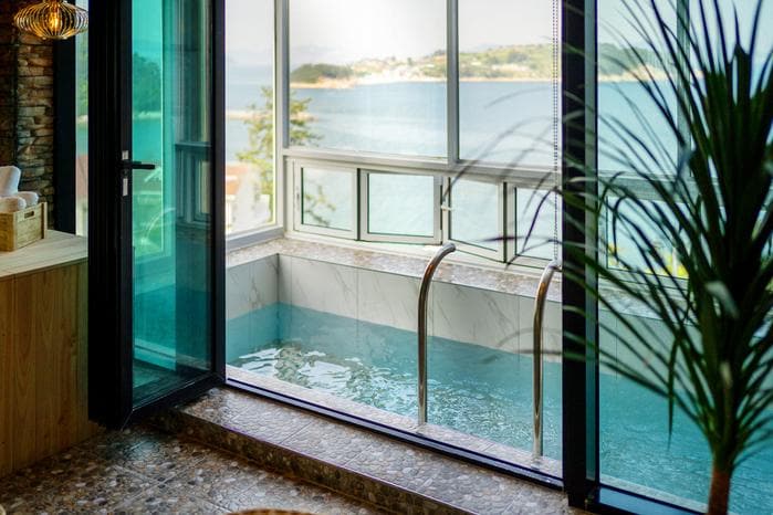 An indoor spa area is featured with large glass doors that open to a view of the sea. A marble spa tub is visible, and sunlight fills the room, highlighting the tranquil atmosphere. Tropical plants add a touch of greenery to the space.