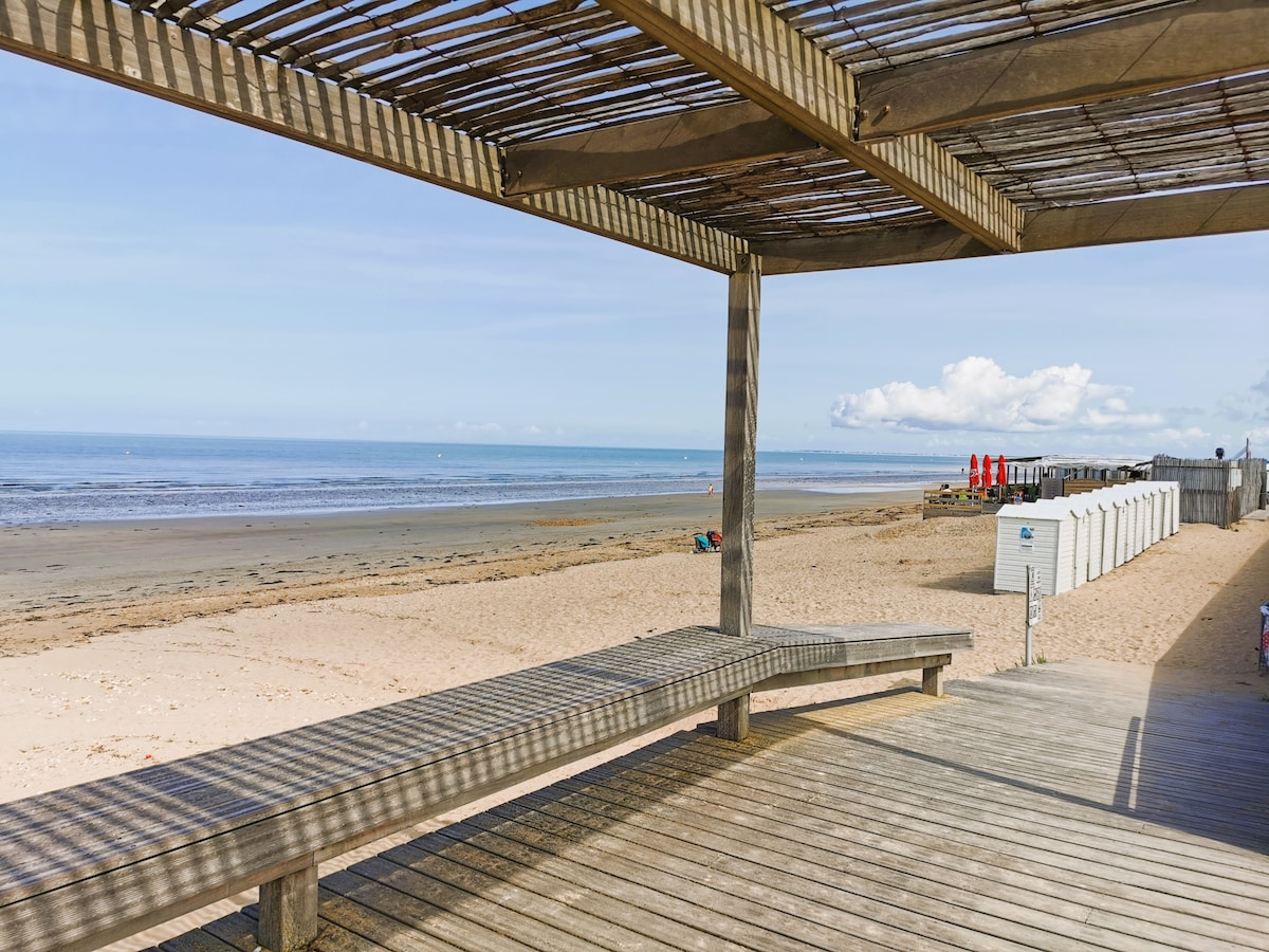 A wooden deck with a shaded structure overlooks a sandy beach and calm sea. Sun loungers are positioned on the sand, and beach cabins line the area in the distance. The sky is clear with scattered clouds, creating a serene beach setting.