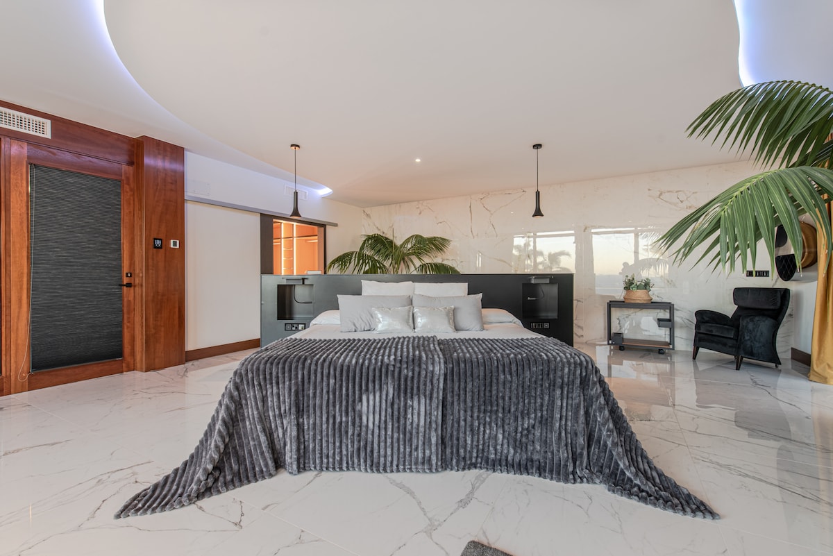 Dormitorio amplio con cama doble, sofá frente a TV, chimenea colgante, vestidor con iluminación LED, ventanales con vistas al mar, escritorio, suelo de mármol, plantas decorativas, cortinas opacas, entrada independiente y zona de estar integrada.