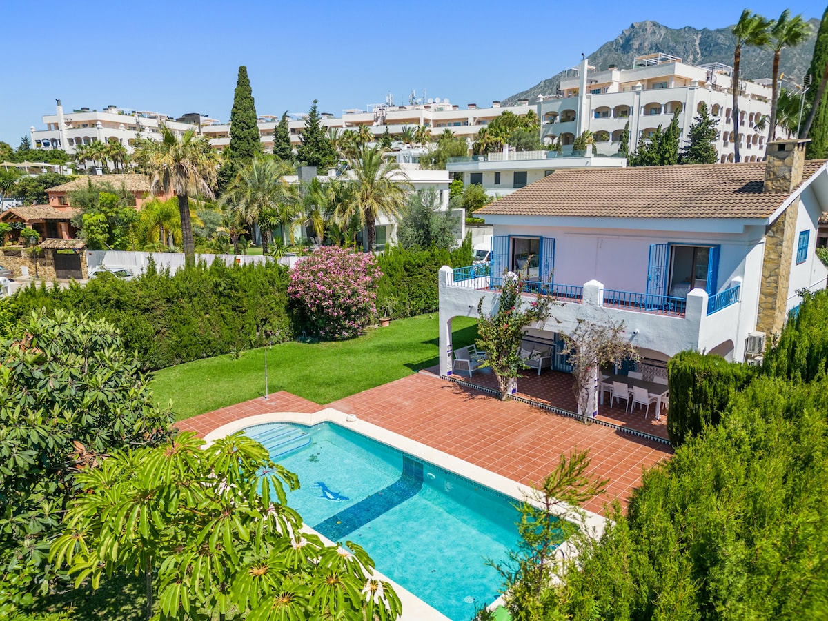 The image showcases a spacious outdoor area featuring a large private swimming pool surrounded by a sun terrace. Lush greenery and colorful flowering plants enhance the garden, while a two-story villa with blue window shutters adds to the inviting atmosphere. The backdrop includes distant buildings and mountains.