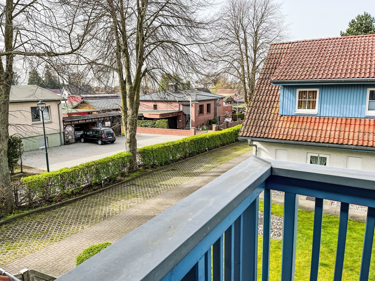 A view from the balcony showcases a peaceful neighborhood scene, framed by leafy trees. The pathway is lined with neatly trimmed hedges, leading to various structures, including parking spaces and residences with varied exterior colors.