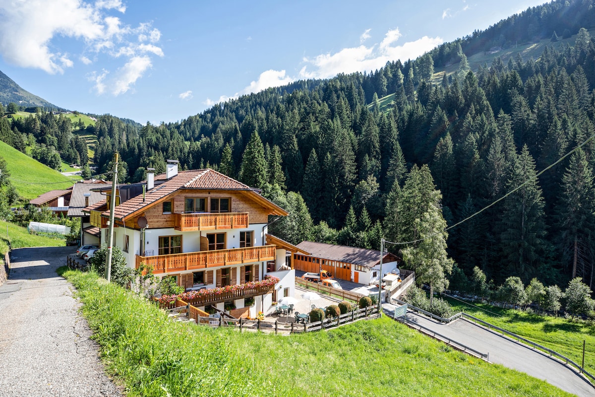 The holiday apartment building is nestled amidst lush greenery and rolling hills, surrounded by forested mountains. The structure features a warm wooden facade and multiple balconies with planters. A well-maintained pathway leads to the entrance, inviting guests into the serene environment.