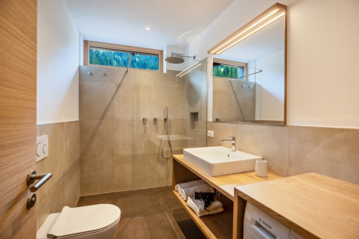 The bathroom features a modern design with a walk-in shower equipped with overhead and handheld fixtures. A spacious vanity with a large mirror and a sink is present, alongside a toilet. Natural light filters in through a window above the shower area.