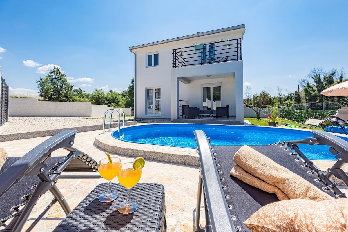 A private pool is surrounded by a sunbathing area featuring comfortable loungers. A table holds two refreshing drinks with garnishes. The villa is located in the background, showcasing a modern design with large windows and a balcony overlooking the tranquil garden and pool area.
