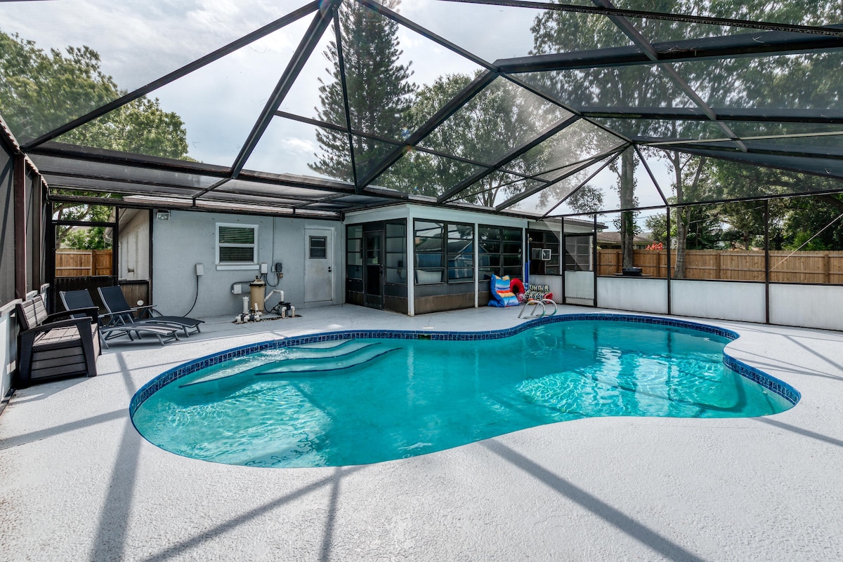 An oversized heated pool is enclosed within a spacious lanai, surrounded by a screened area. A few lounger chairs are positioned nearby, while the pool features a gentle curve. Natural light filters through the mesh roof, creating a bright, inviting atmosphere for relaxation.