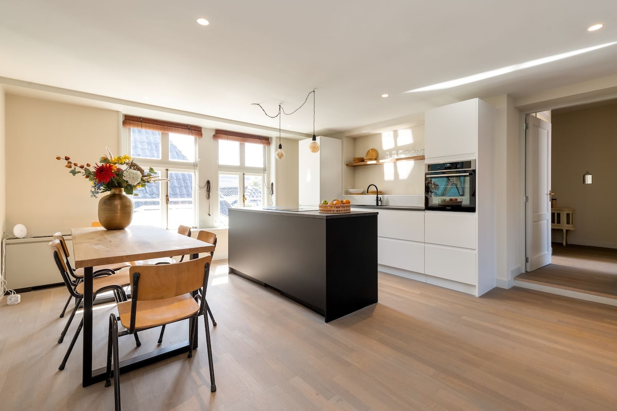 A modern kitchen space is highlighted by an island with a black countertop and a fitted oven. A dining table is surrounded by wooden chairs, adorned with a vase of fresh flowers. Natural light streams through multiple windows, creating a bright and airy environment.