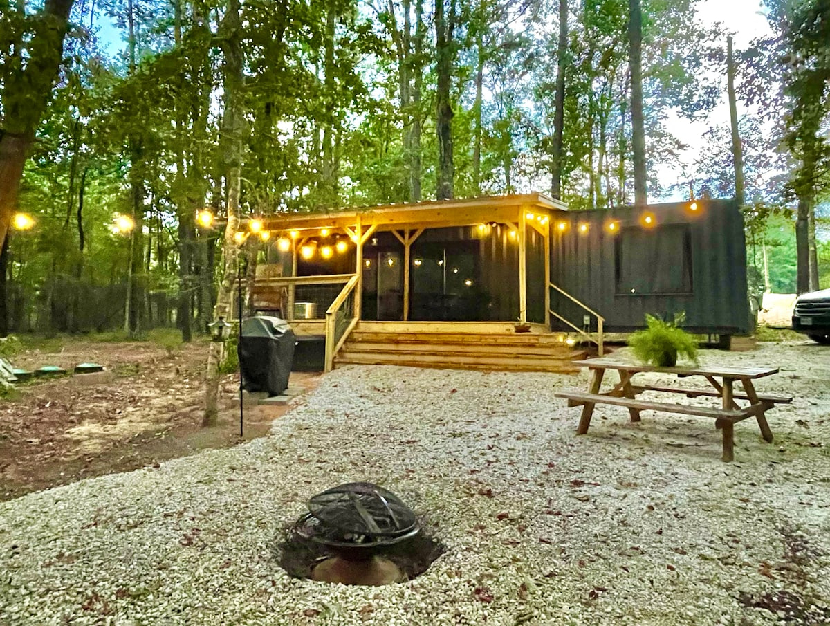 A cozy cabin is set amidst a wooded area, featuring a wooden deck with string lights. A fire pit is located on a gravel path in the foreground, while a picnic table provides an area for outdoor dining. Tall trees surround the space.