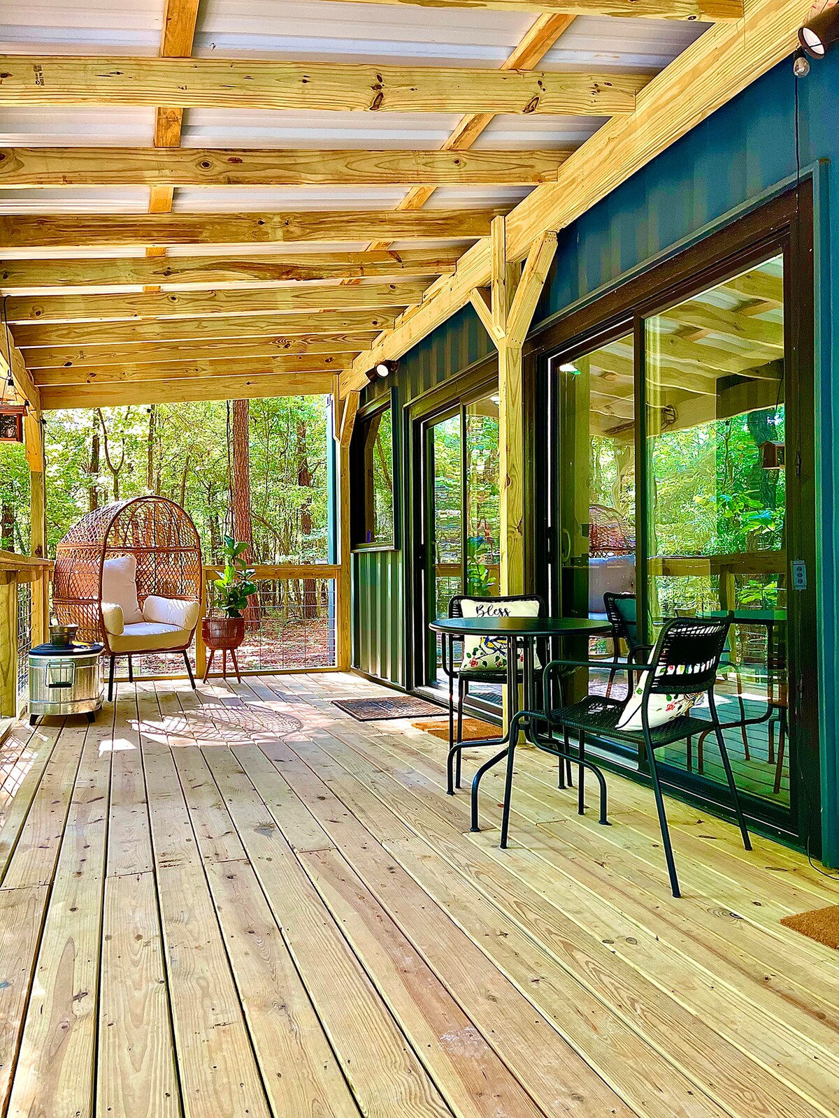 A spacious outdoor deck is surrounded by tall trees, featuring a comfortable hanging chair and a small table with two chairs. Glass doors provide easy access to the interior, enhancing the connection with nature.