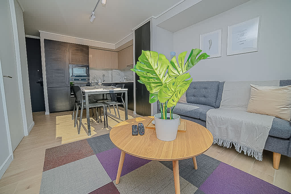 A living area features a round wooden table with a potted plant at its center. Gray sofas are arranged with a throw blanket draped casually. The kitchenette is visible in the background, with a dining table set for four. Light-colored flooring enhances the space.