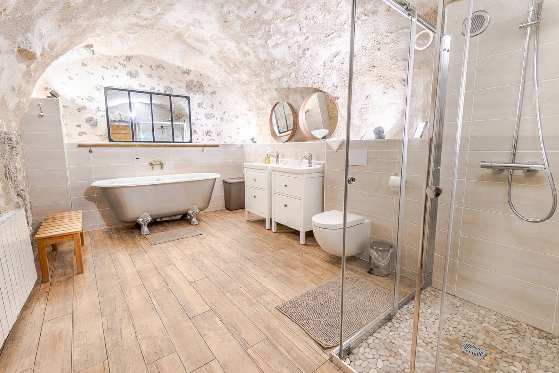 A spacious bathroom features a freestanding bathtub and a modern shower area with glass doors. Two circular mirrors above a double vanity sink add elegance. Natural light is filtered through a large window, highlighting the stone ceiling and warm wooden flooring.
