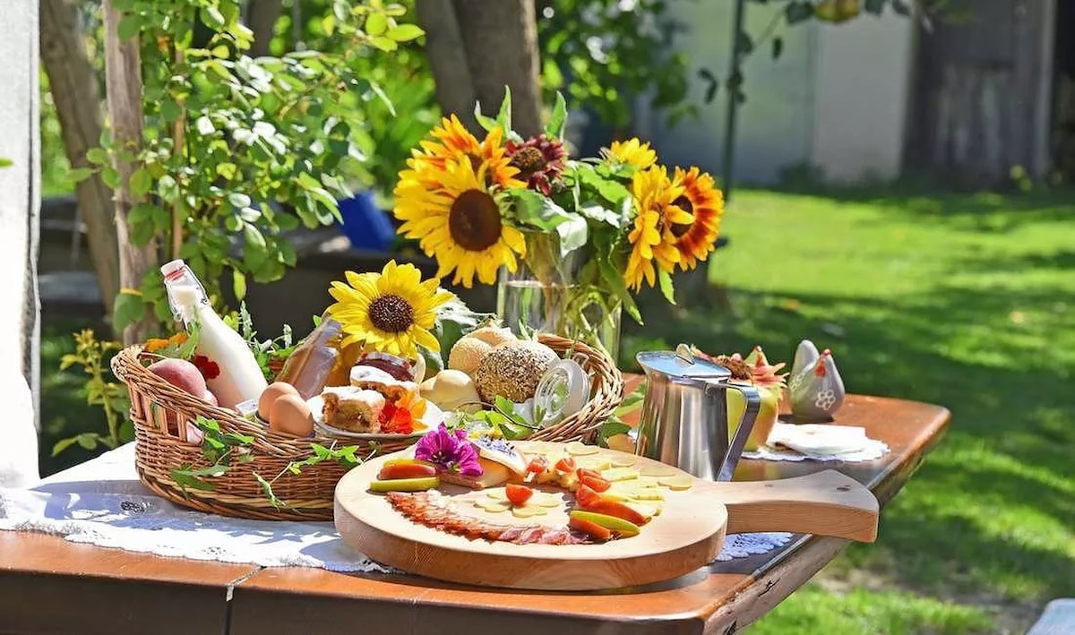 A table is set outdoors, showcasing a variety of food and drinks. A wooden platter holds assorted slices of fruit and pastries, while a woven basket contains additional breads and snacks. Sunflowers and greenery create a vibrant natural backdrop in the outdoor space.