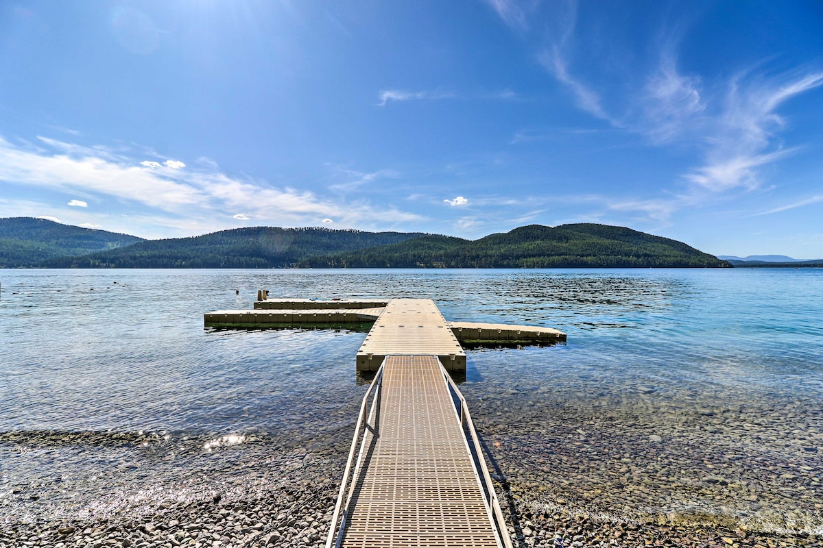 A wooden dock extends over the clear waters of Whitefish Lake, inviting guests to enjoy views of surrounding hills and trees. The shoreline features smooth pebbles, while gentle waves create a serene atmosphere under a sunny blue sky.