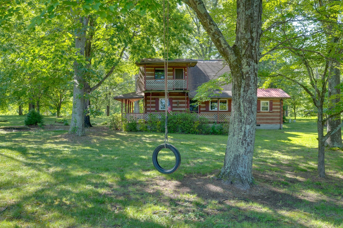 A serene outdoor setting showcases a cabin surrounded by lush greenery. A tire swing is suspended from a tree, complemented by a spacious yard. The cabin features two stories, with a porch offering ample seating and large windows that invite natural light.