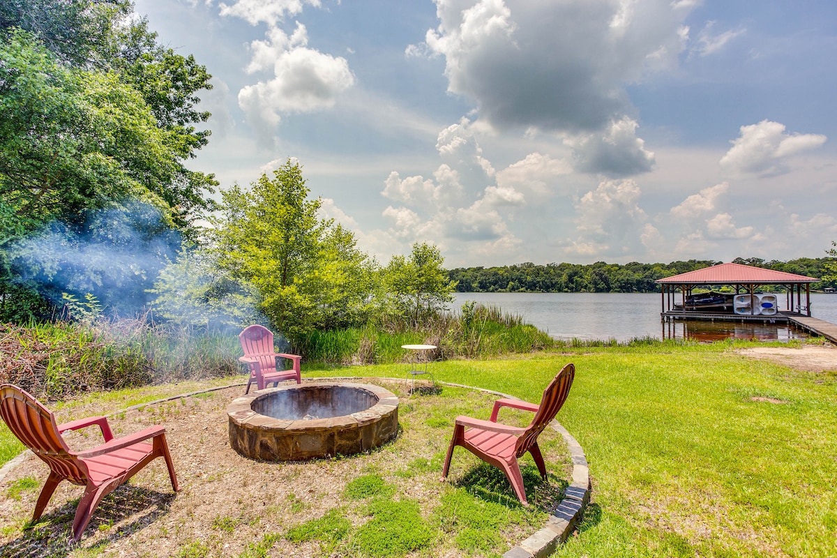 A tranquil outdoor space features a stone fire pit surrounded by four red chairs, set against a backdrop of lush greenery and a peaceful lake view. A covered dock extends into the water, under a partly cloudy sky.