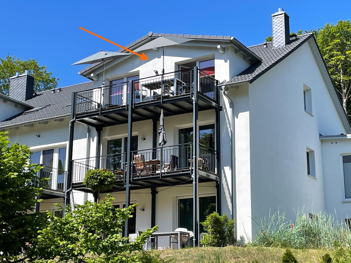 The exterior of a spacious two-story building is visible, featuring a furnished balcony with seating. The facade is adorned with large windows, allowing natural light to flow inside. Surrounding greenery complements the tranquil atmosphere.