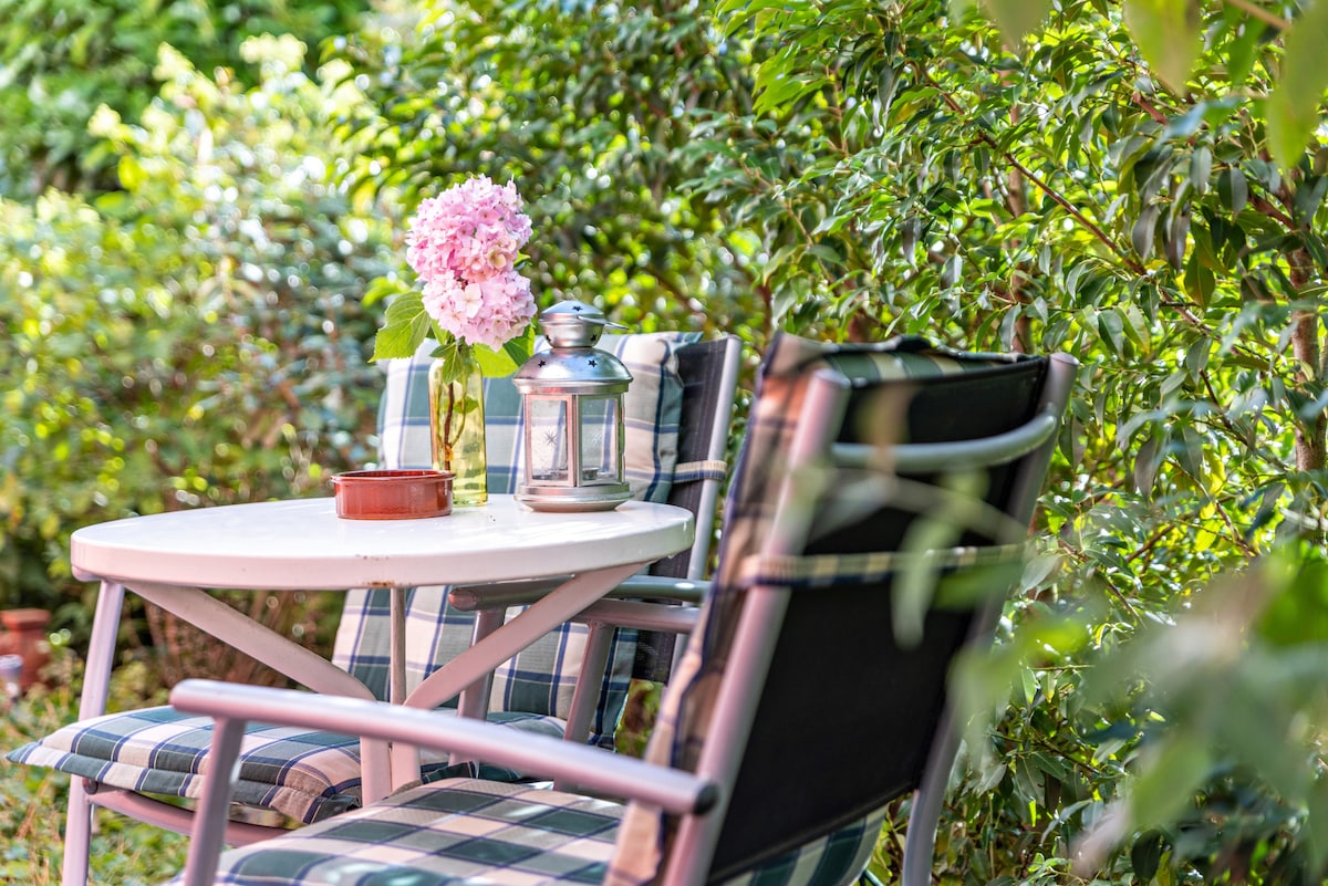 A small outdoor seating area is surrounded by lush greenery. A round table with a vase of pink flowers sits in the center, accompanied by two cushioned chairs. A decorative lantern is placed on the table, creating a serene, natural ambiance.