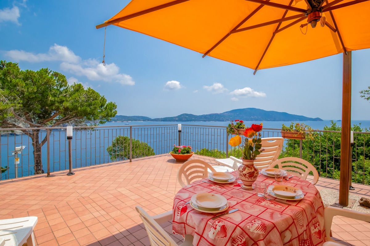 An outdoor terrace features a round table set for four, adorned with a floral centerpiece. A large orange umbrella provides shade, while the stunning view of the sea and nearby hills creates a serene backdrop. The tiled surface is highlighted by vibrant colors.