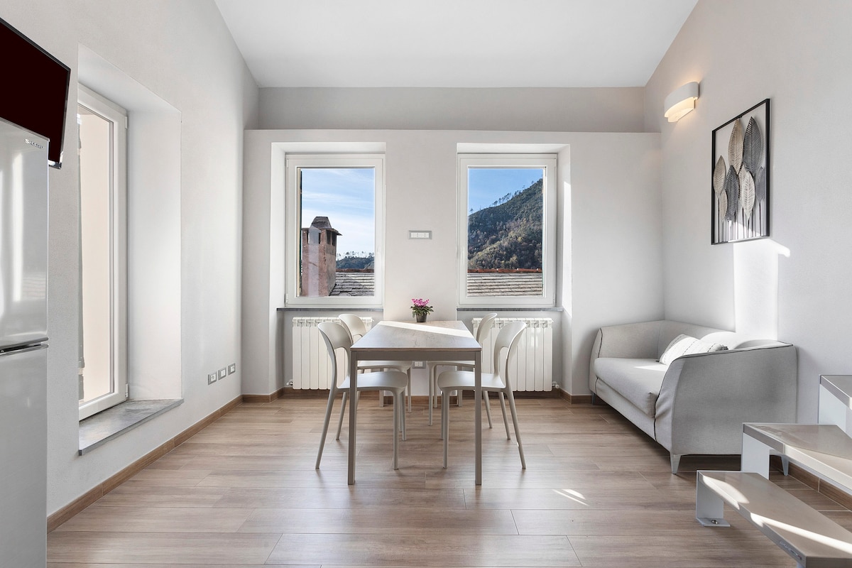 A light and airy living space is depicted, featuring a dining table surrounded by four chairs. Large windows provide natural light and reveal views of the outside landscape. A sofa is positioned against the wall, complementing the neutral color scheme of the room.