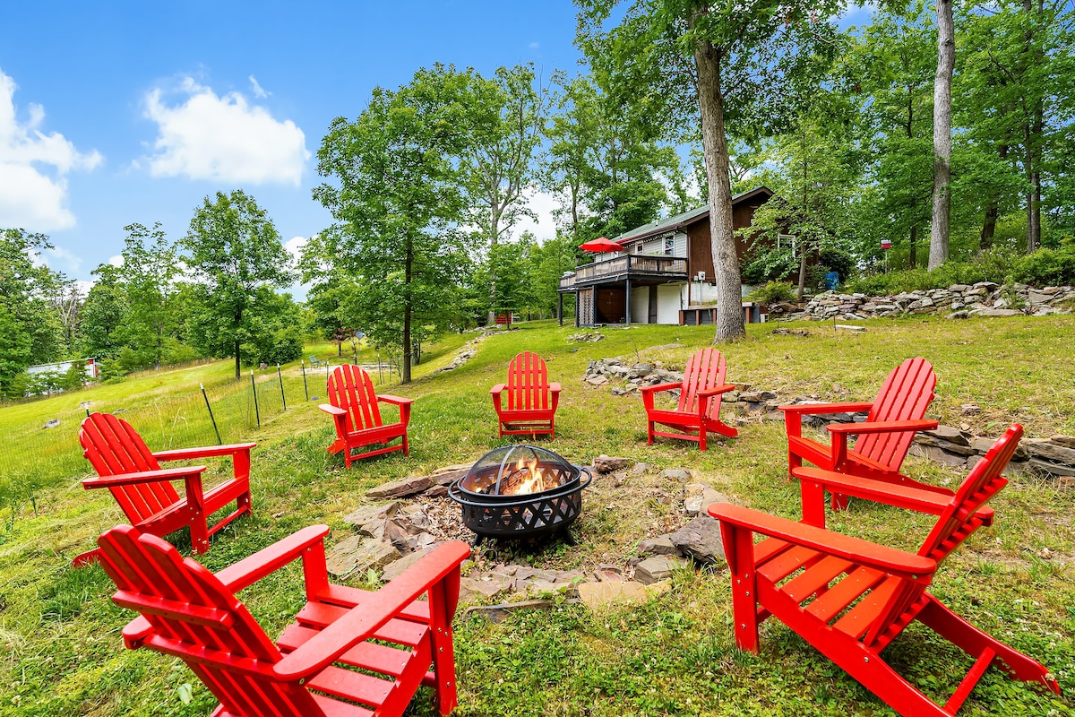 An outdoor fire pit area features several red Adirondack chairs arranged in a circle, inviting conversation and relaxation. Lush greenery surrounds the space, complemented by the cabin in the background, creating a serene outdoor environment for gatherings.