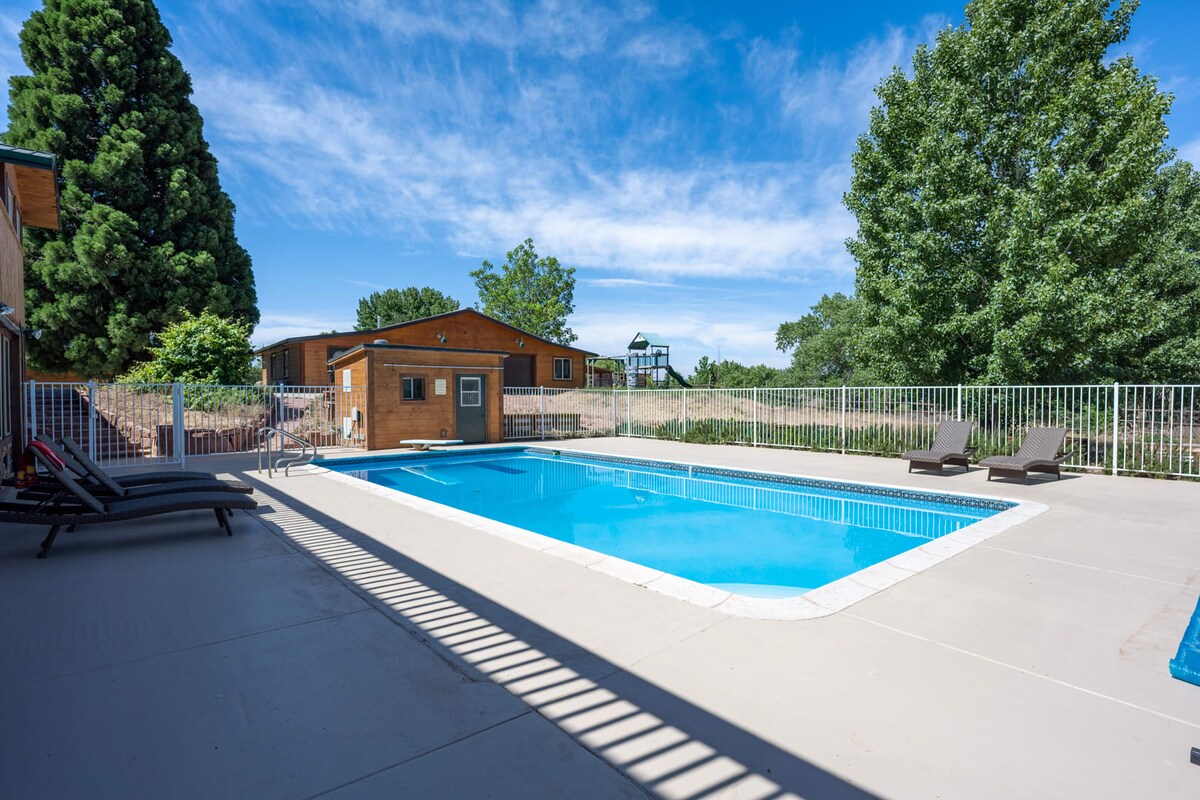 A spacious heated pool is surrounded by a manicured patio area featuring lounge chairs. A securely fenced perimeter ensures safety. Lush trees and greenery provide a pleasant backdrop, while a playground can be seen in the distance, contributing to a family-friendly atmosphere.