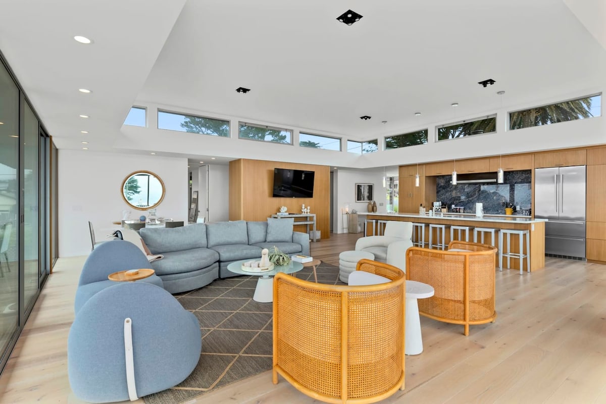 An open-concept living area showcases a soft blue sectional sofa alongside two modern white chairs and a round wooden table. The kitchen, equipped with wood cabinets and stainless-steel appliances, extends into the space, providing a seamless flow. Large windows offer views of the outside.