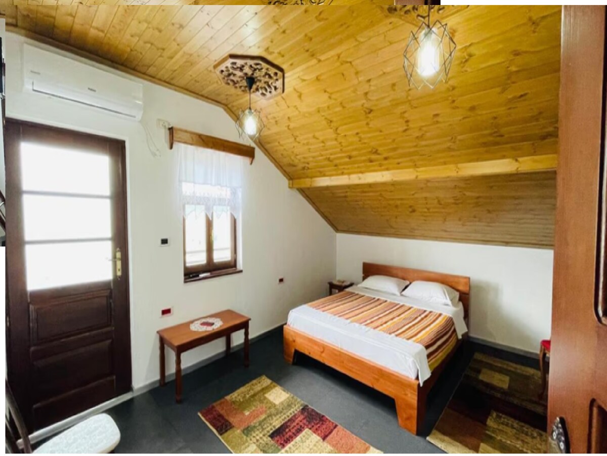 A room features a wooden ceiling with exposed beams and two pendant lights. A spacious bed with a multi-colored blanket is positioned against the wall, complemented by a small wooden table. Natural light enters through a window, and a door leads to the outside.
