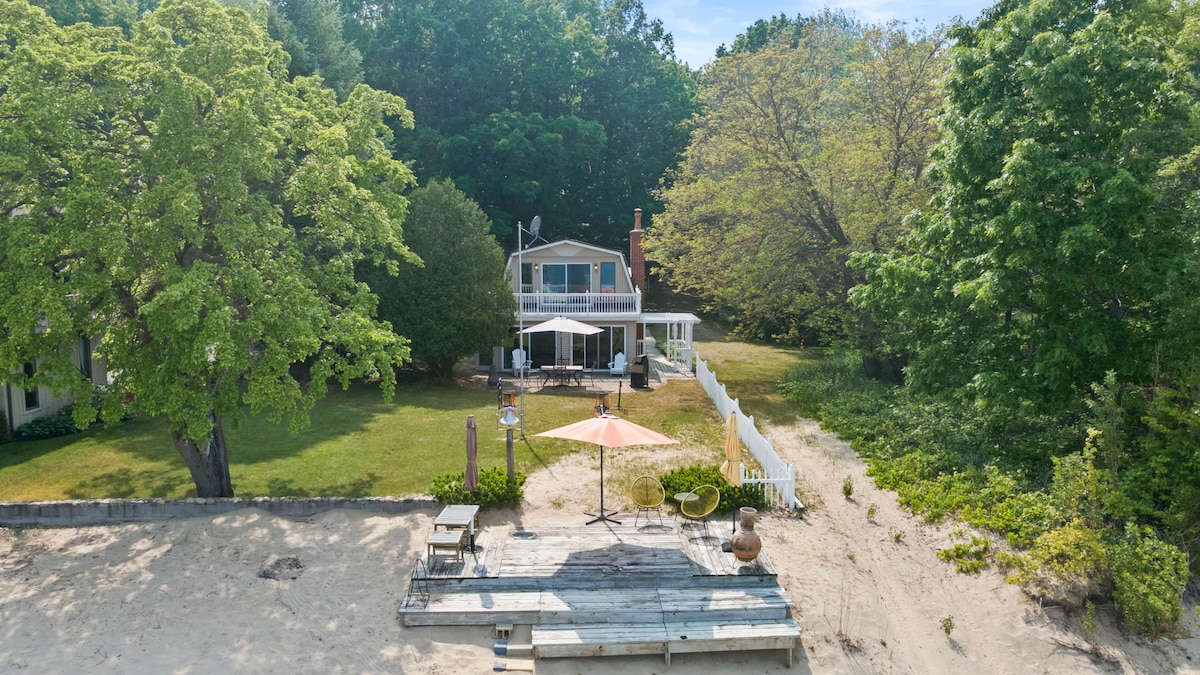 A two-story beachfront retreat is surrounded by lush greenery, featuring a spacious yard. A patio with an umbrella and outdoor seating is visible in front, while large windows allow for natural light to illuminate the interior.