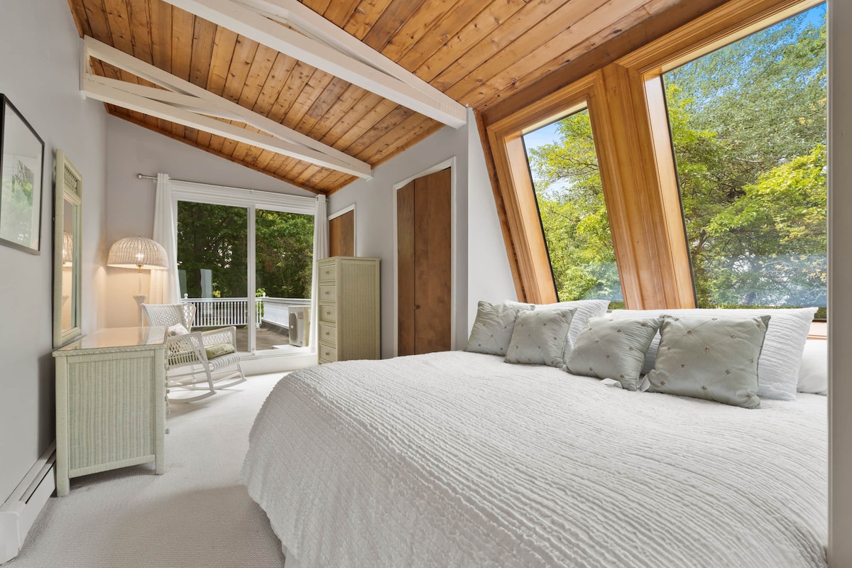 A light-filled bedroom features large, angled windows that offer views of lush greenery. A spacious bed with a textured white coverlet is complemented by decorative pillows. A wicker chair and dresser enhance the room's inviting aesthetic, while natural wood beams add character to the ceiling.