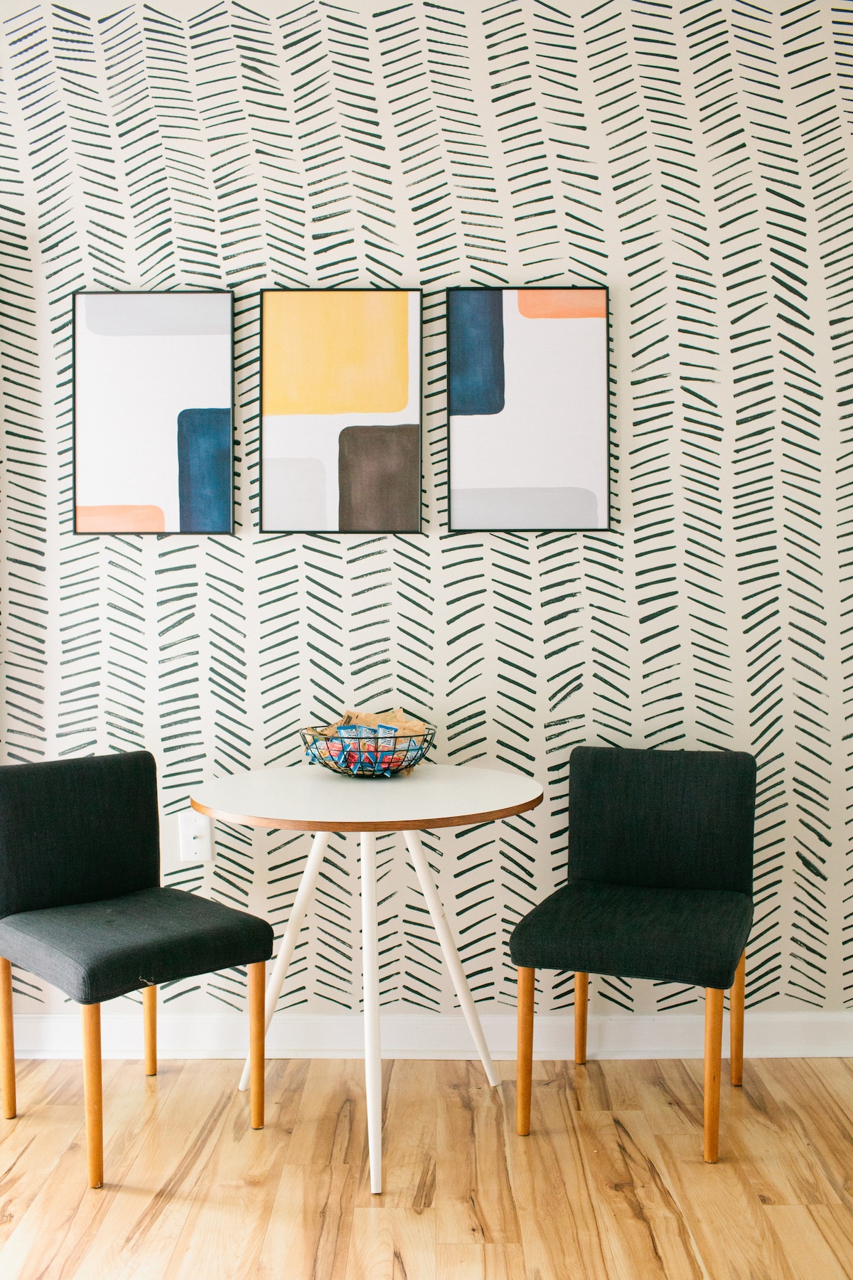 A dining area features a round white table surrounded by two dark chairs. Above the table, three framed artworks are displayed on a patterned wall, which showcases a contemporary design. A colorful bowl sits at the center of the table.