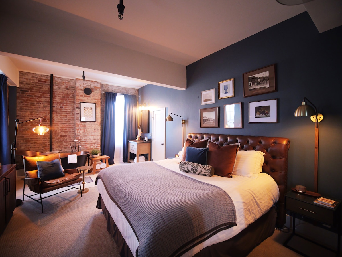 A spacious guest room features exposed brick walls and a large window with blue curtains. A plush king bed with decorative pillows is complemented by bedside tables and stylish lamps. A seating area with a wooden chair adds comfort and warmth to the space.