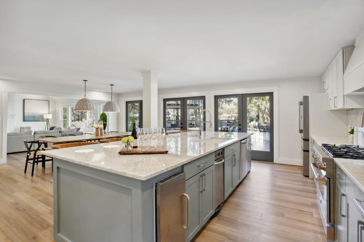 An open-concept kitchen is highlighted by a large island featuring a light-colored countertop. Modern appliances are visible, complemented by light cabinetry. Large glass doors provide a view of the outdoor area, allowing natural light to fill the space. Seating options are available at the island.