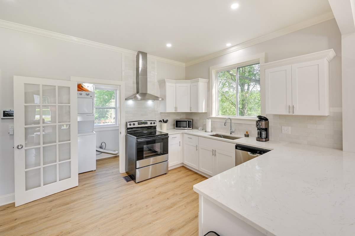 A modern kitchen design features white cabinetry and a spacious countertop. Stainless steel appliances, including an oven and microwave, are present. A window offers natural light and views of the outdoors, while a pantry is visible behind glass doors.