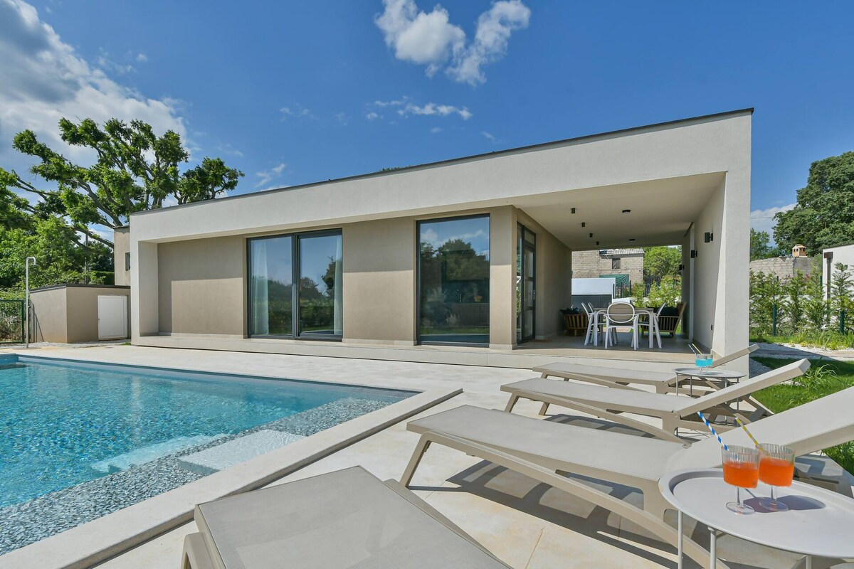 A modern house is positioned near a tranquil pool, featuring large glass doors that open to a sunlit living area. Comfortable lounge chairs are arranged by the poolside, with a small table displaying two refreshing drinks. Lush greenery surrounds the property.