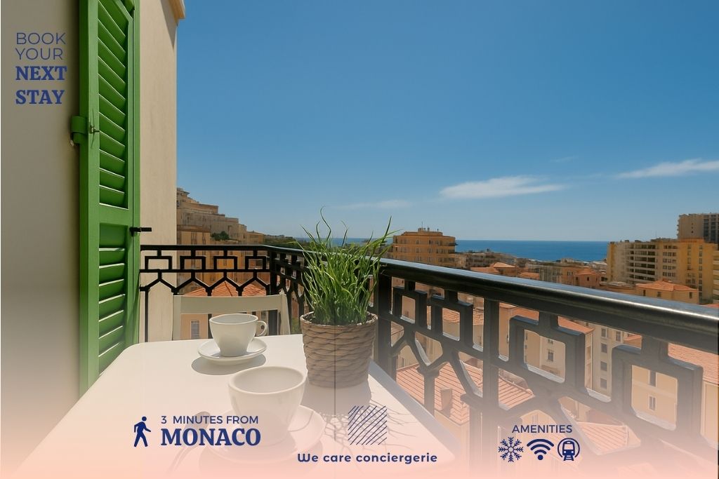 A balcony table is set with two cups and a potted plant, framed by a decorative railing. The expansive view reveals the rooftops of Monaco and the serene sea under a clear blue sky.