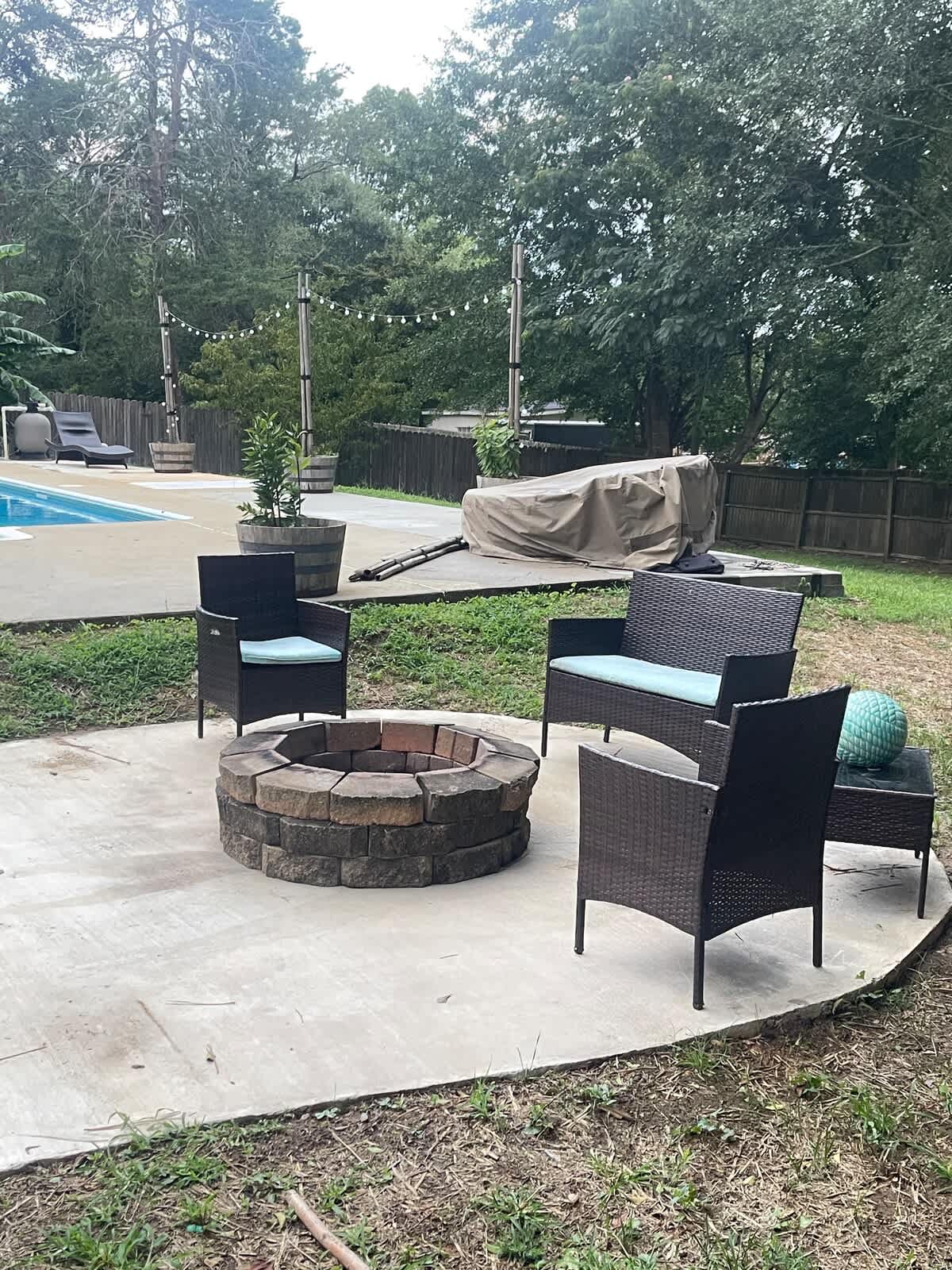 A circular firepit, surrounded by five woven chairs with cushions, is positioned on a concrete patio. In the background, an in-ground pool is visible, accompanied by chair loungers and a canopy cover. Natural greenery and trees frame the relaxing outdoor area.