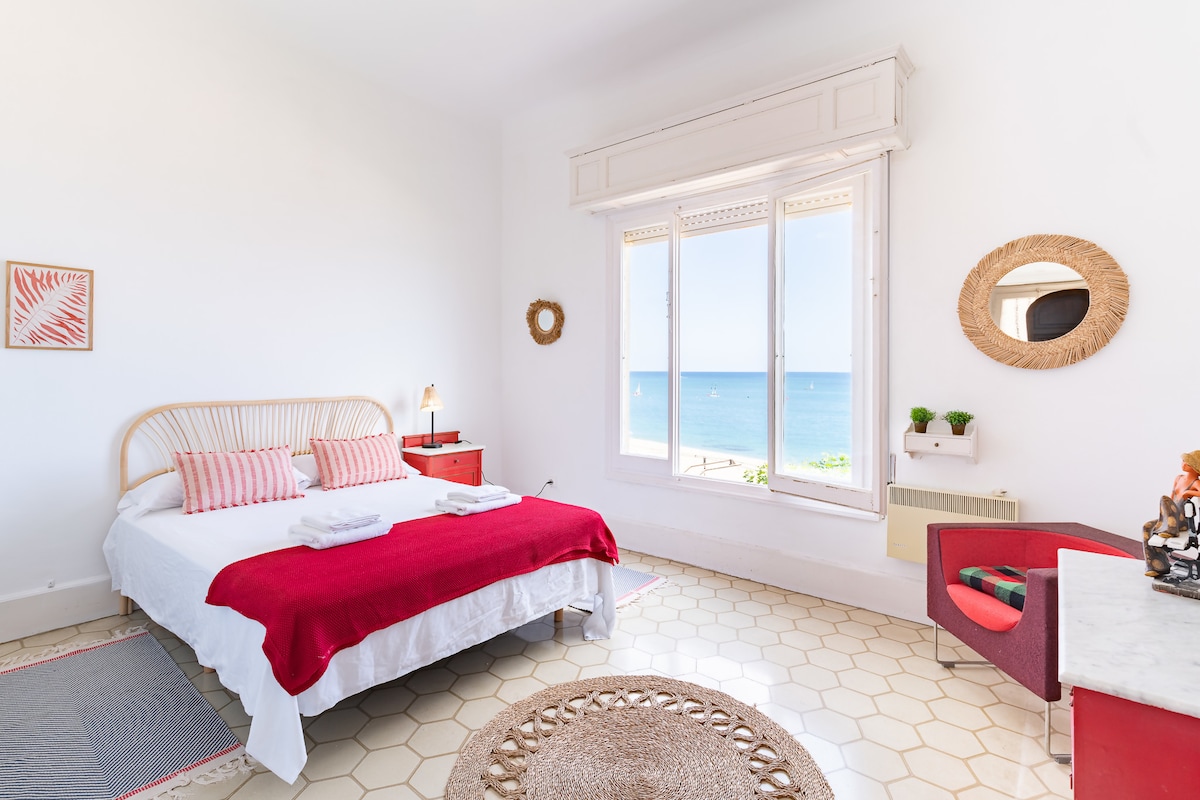 A bright bedroom is showcased with large windows offering ocean views. The spacious bed is adorned with white linens and red accents, accompanied by a small nightstand. A cozy seating area features a red chair and a soft rug on the tiled floor.