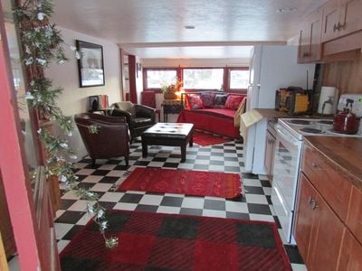 The open living area features a checkered floor in black and white, complemented by vibrant red area rugs. Cozy seating includes two leather chairs and a fabric couch. A kitchenette is visible, equipped with basic appliances. Large windows allow natural light to illuminate the space.