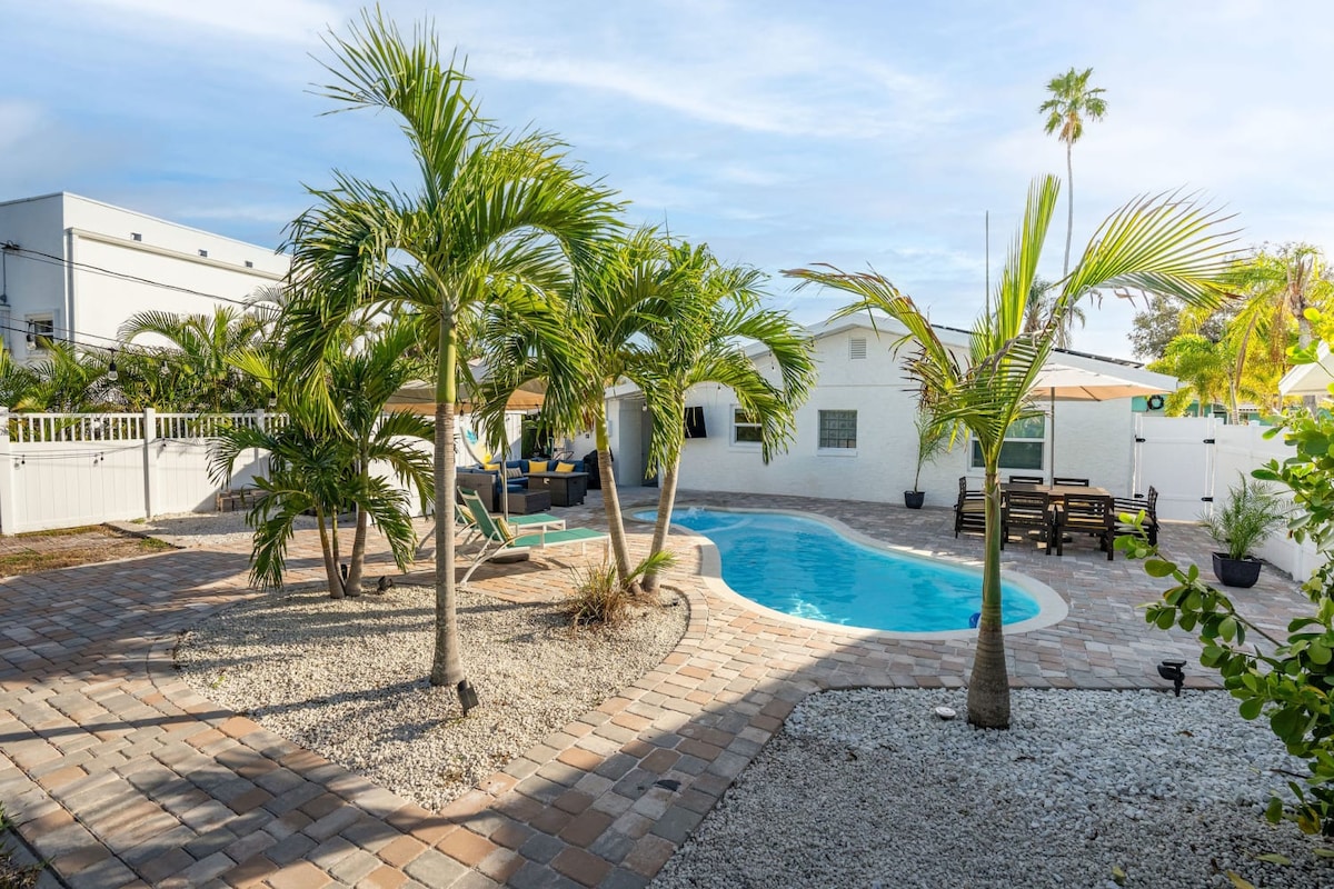 A serene outdoor area features a glistening saltwater pool surrounded by tropical landscaping. Lush palm trees provide shade, while a dining area with an umbrella and comfortable seating invites relaxation. A firepit and lounge chairs complete the tranquil backyard oasis.