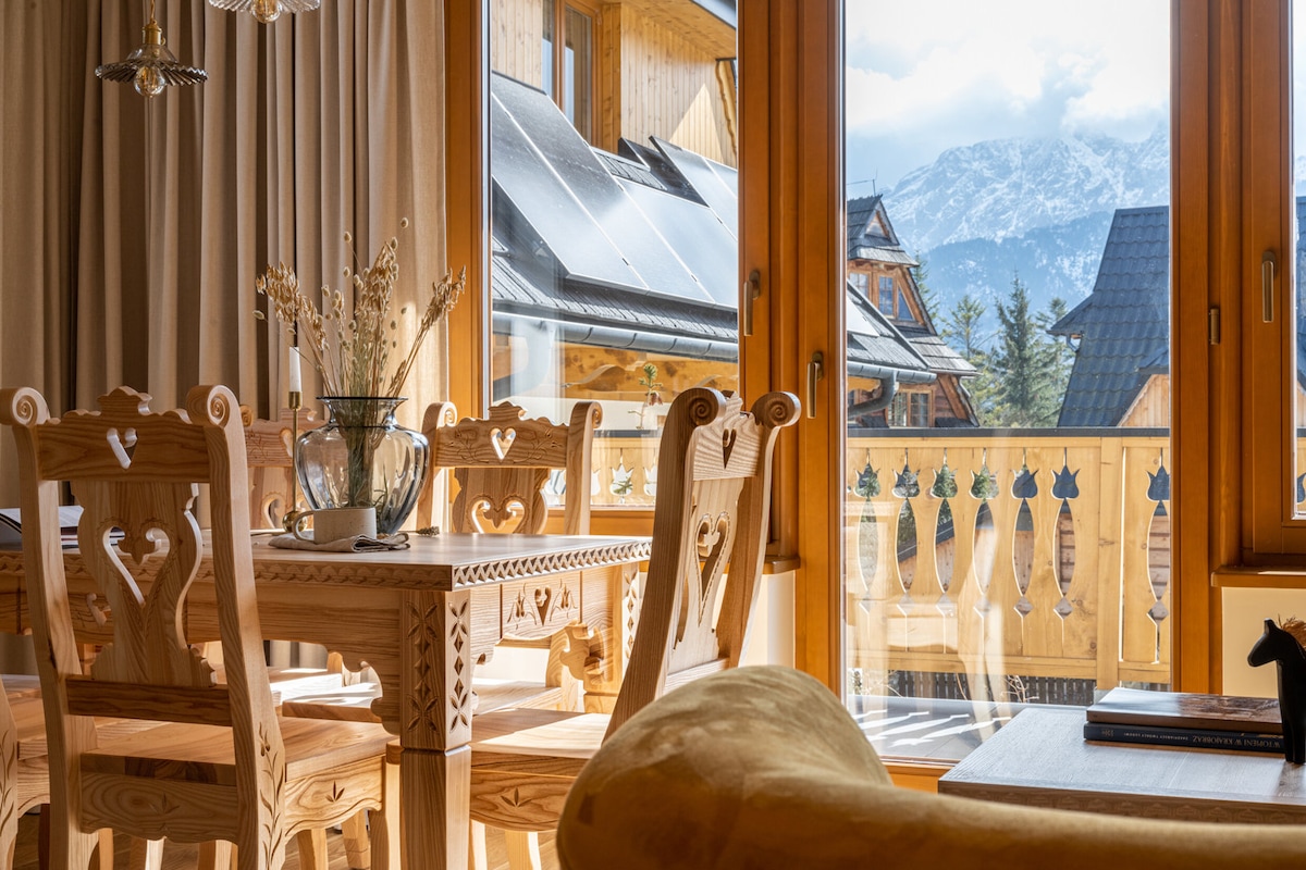 A wooden dining table is surrounded by intricately carved chairs, creating a warm dining area. Large windows provide a view of the mountains, while soft natural light enhances the inviting atmosphere of the space.