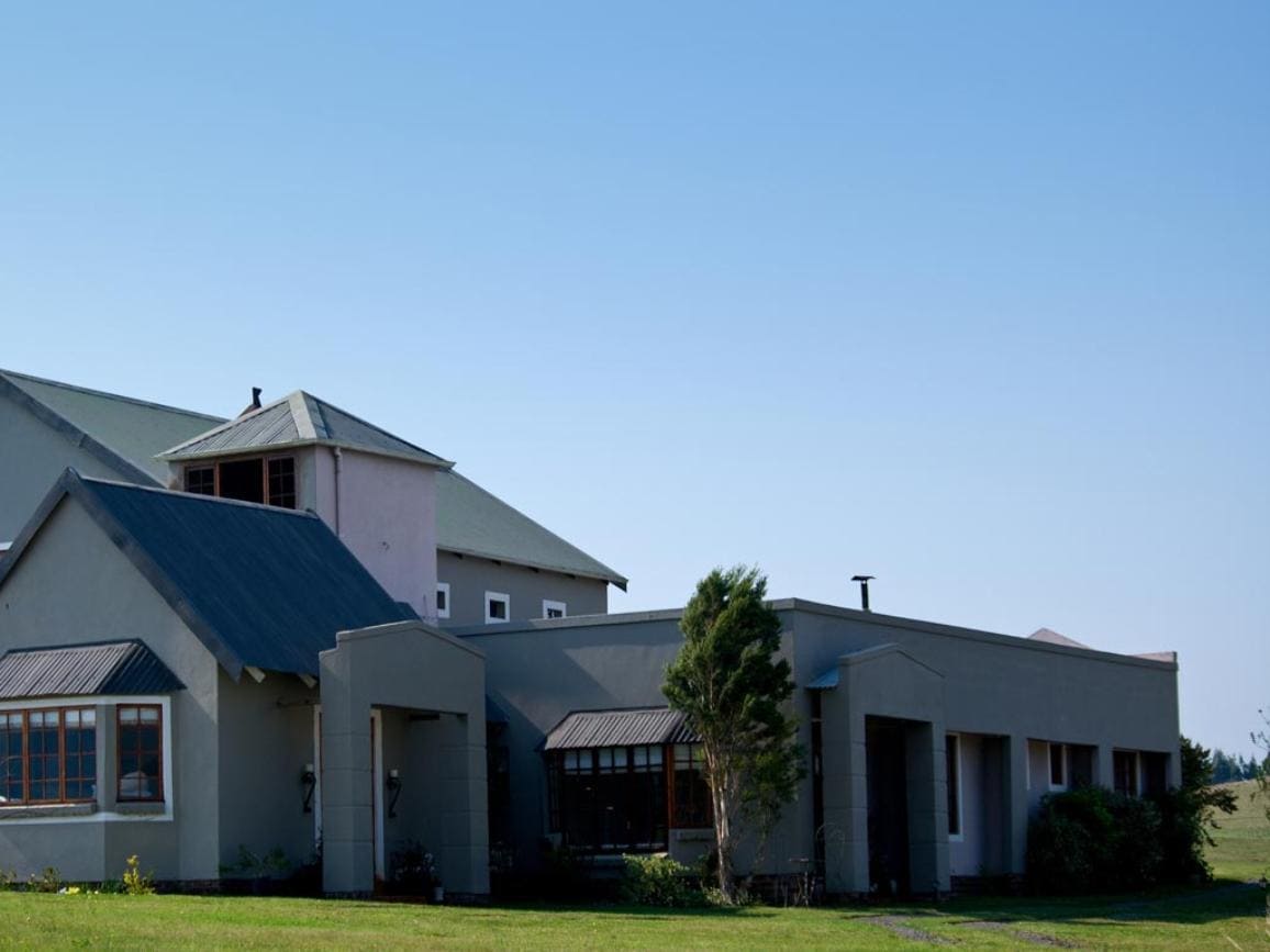 The exterior of the cottage features a mix of colors, with a prominent grey wall and a sloped roof. Large windows provide natural light, while a lawn surrounds the building, enhancing the outdoor space. The structure’s multi-level design adds visual interest.