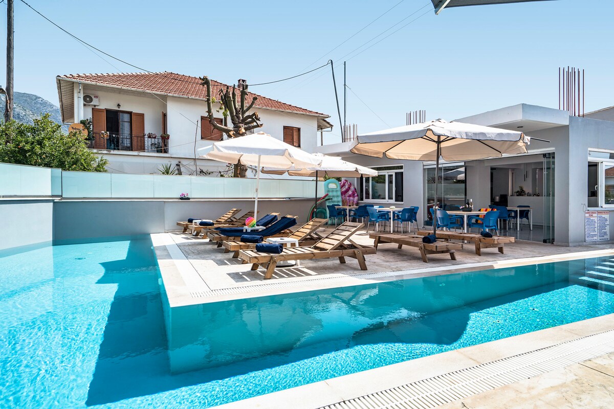 A serene outdoor area is showcased, featuring a clear blue pool surrounded by lounge chairs. Sun umbrellas provide shade, while a comfortable seating area is visible nearby. The backdrop includes a modern building, contributing to a relaxed atmosphere ideal for leisure.