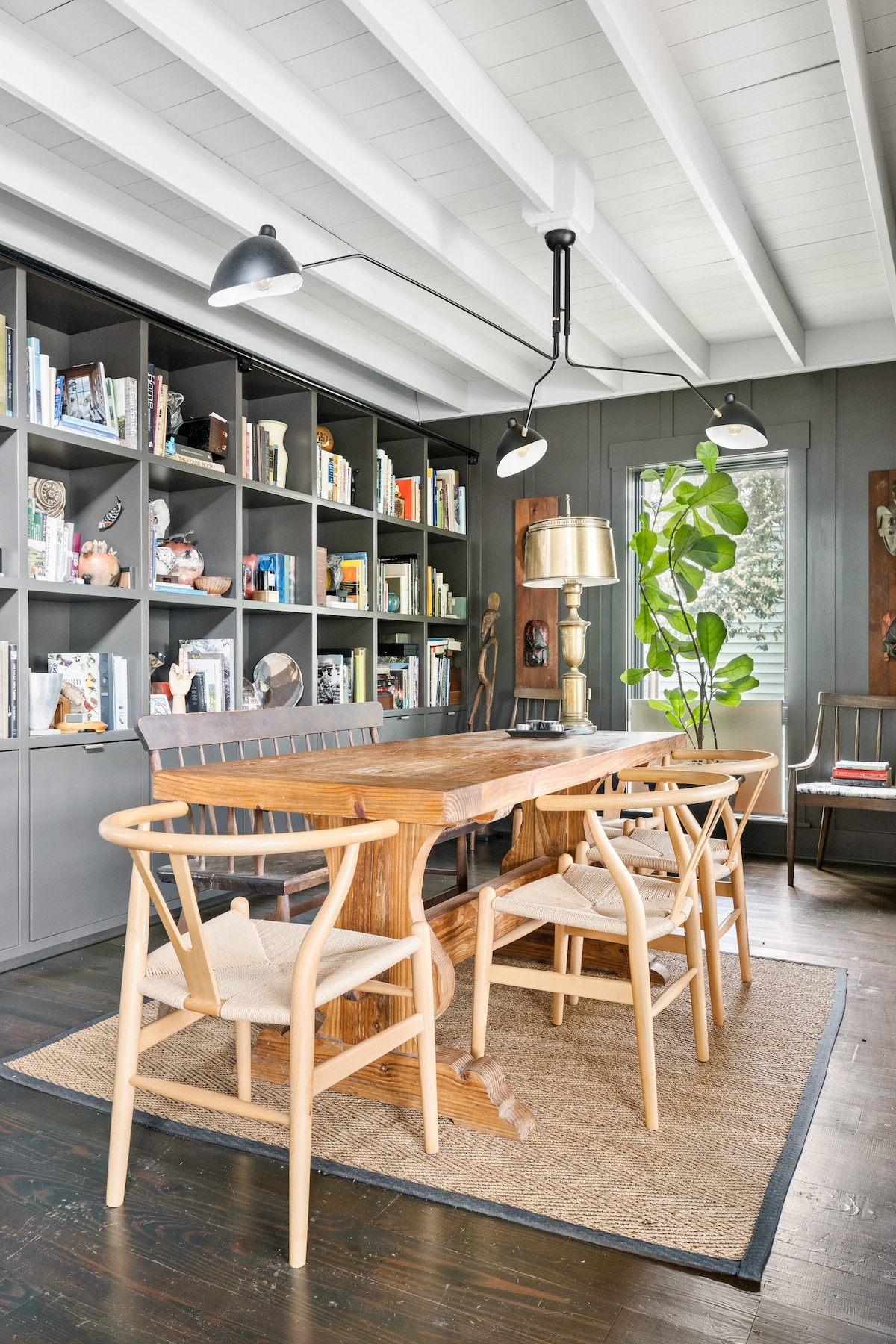A dining area features a natural wood table surrounded by four light-colored chairs. A shelving unit filled with books and decorative items lines the wall, complemented by a plant and a floor lamp. Recessed lighting illuminates the space with a warm glow.